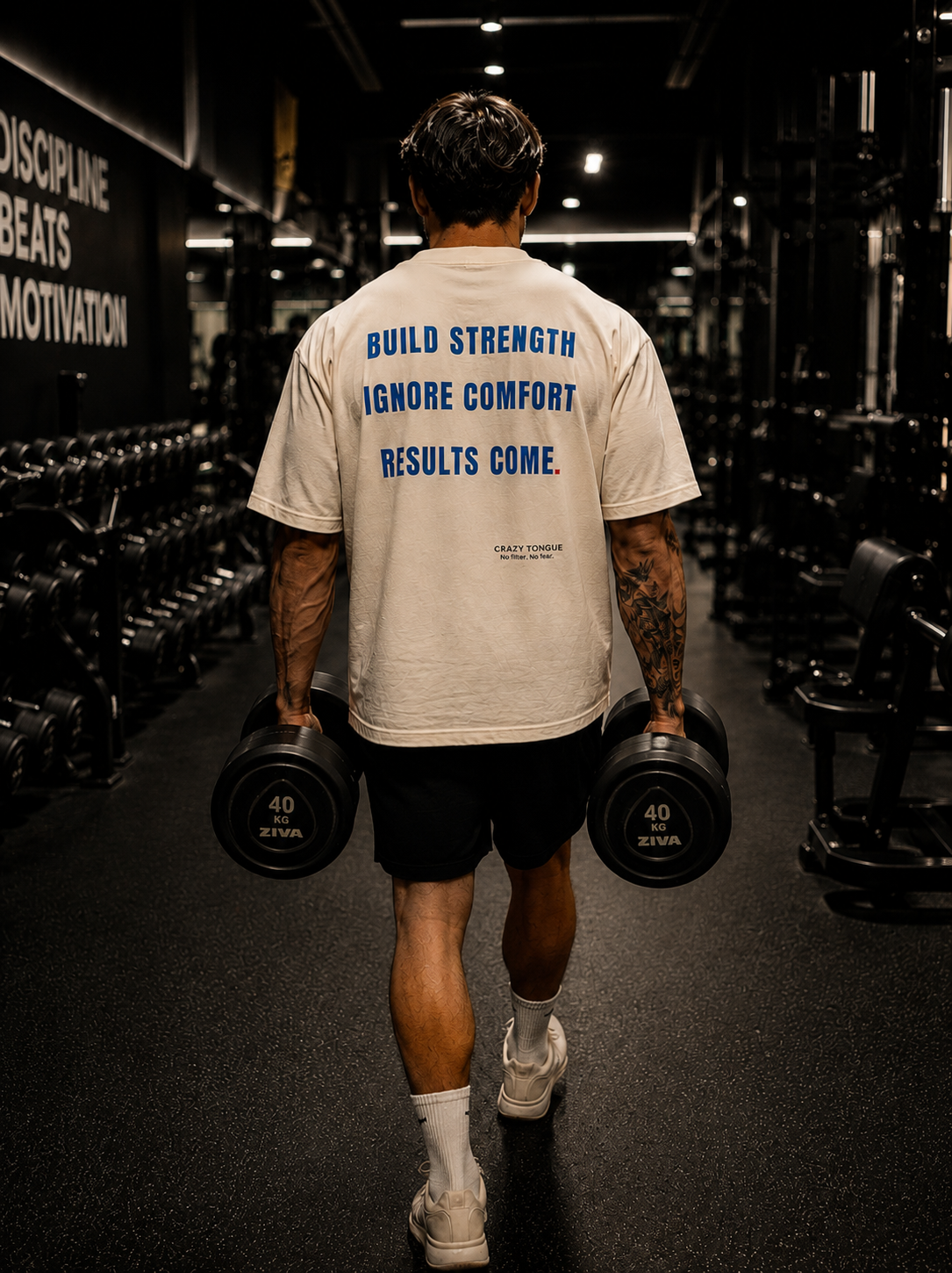 Homme portant le t-shirt crème Crazy Tongue BUILD STRENGTH de dos dans une salle de sport.